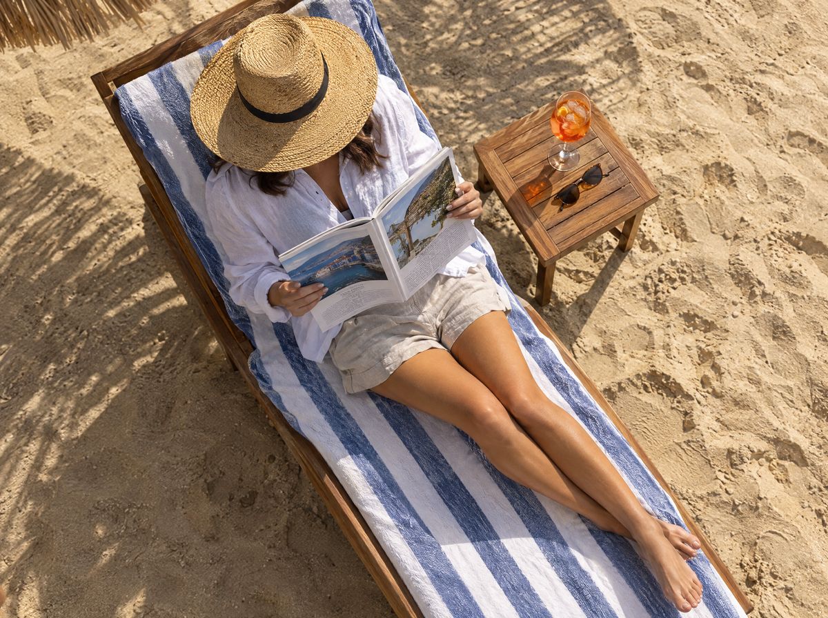 Woman on a beach lounger reading a magazine, with a cocktail on the small table beside her